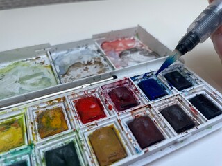 Paintbrush with blue paint getting color from messy watercolor palette 