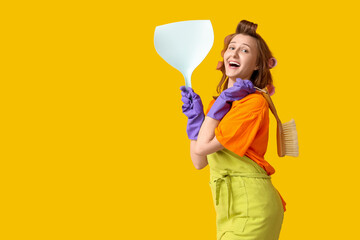 Young woman with hair curlers holding dustpan and brush on yellow background