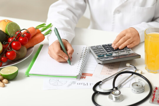 Young female nutritionist with healthy food, glass of juice and calculator writing diet plan in office, closeup