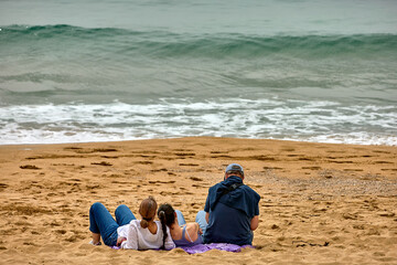 family sits on a sandy beach facing the ocean, waves rolling in under an overcast sky  footprints scattered across the shore Palma Mallorca, Spain - March 20, 2025A © Goodwoon Mallorca
