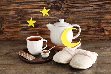 Cup and teapot of tea with bowl of honey, pills, slippers, stars and moon made of paper on wooden background