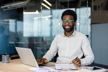 Obraz premium A happy, young professional works on a laptop, makes notes, and reviews data at his desk in a modern office with glass walls.