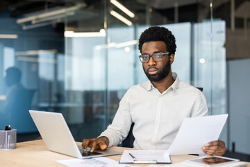 A focused professional balances reviewing documents and working on his laptop in a modern office...