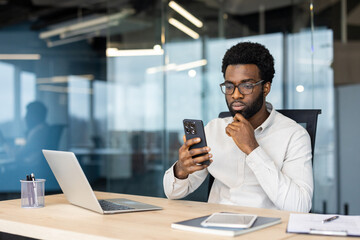 A serious, young professional man in glasses checks his smartphone while sitting at his desk in a modern office.