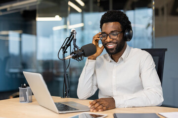 Smiling African American man wearing headphones, sitting at a desk with a microphone and laptop...
