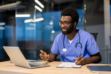 A black male doctor in scrubs and stethoscope smiles while taking notes during a telehealth...