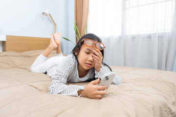Asian Thai Little girl is lying on the bed with using smartphone, Wearing sunglasses and Eye strain and eye rubbing, Visual health