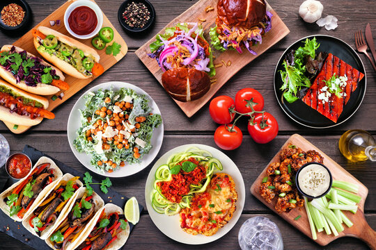 Healthy plant based vegetarian food table scene. Top down view on a dark wood background. Jackfruit burger, mushroom tacos, watermelon and cauliflower steaks, carrot hot dogs, walnut bolognese
