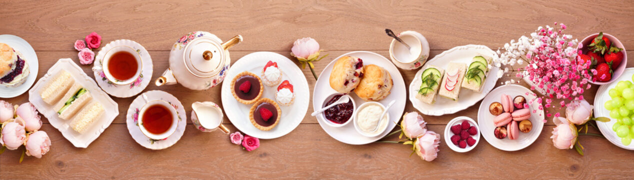 Afternoon tea table scene against a light brown wood banner background. Vintage tea set, finger sandwiches, scones, and desserts. Overhead view.