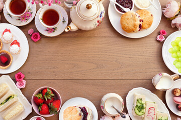 Afternoon tea frame against a light brown wood banner background. Vintage tea set, finger sandwiches, scones, and desserts. Top down view with copy space.
