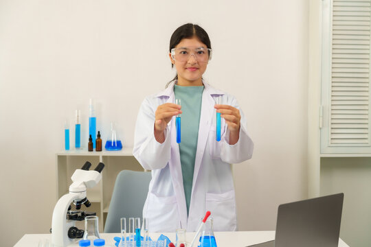 Teenage Caucasian schoolgirl wearing lab coat and protective glasses holding test tubes filled with blue liquid while conducting science experiment during secondary school chemistry class