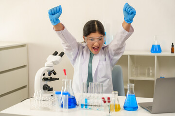 Teenage Caucasian girl wearing blue gloves and laboratory coat raising arms in excitement celebrating experiment success while smiling cheerfully near beakers test tubes and microscope on desk