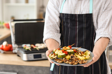 Young man with plate of tasty cooked vegetables near modern electric grill in kitchen, closeup