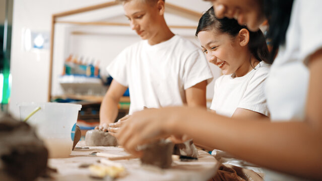 Handsome student dipping hand in to water to soften clay at art classroom. Group of diverse children working or modeling vase. Happy boy put water to young girl while laugh with happy. Edification.