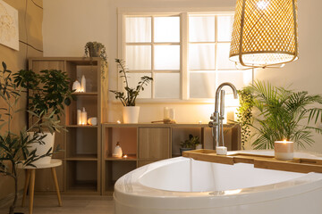 Interior of stylish bathroom with bathtub, shelf unit and glowing lamp in evening
