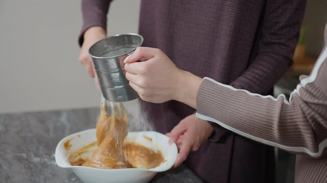 Partial view of girl helping her aunt in kitchen, holding metal sifter while aunt stirs batter in bow, they work together in bright, modern kitchen