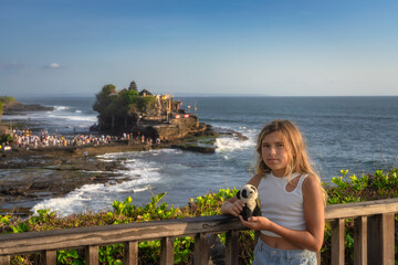 A cheerful girl poses with her toy panda at Balis Tanah Lot Temple, surrounded by stunning ocean views and excited tourists, creating a perfect holiday memory, Bali, Indonesia