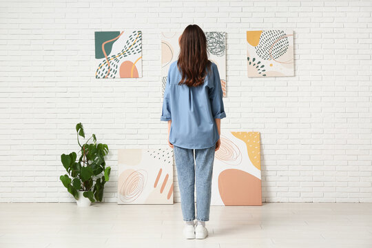 Young woman looking at paintings on white brick wall in art gallery, back view
