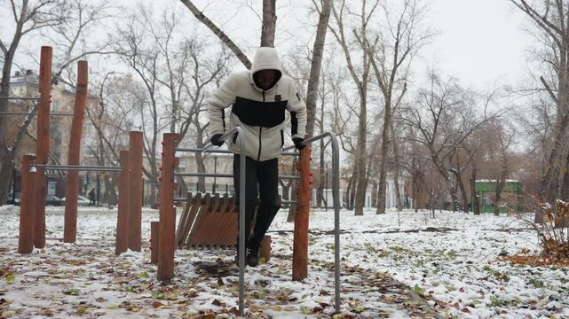 Young adult performs muscle-building workout on parallel bars in snowy outdoor park, dressed in warm winter clothing, he maintains balance and strength while exercising amidst a cold environment