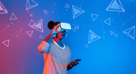 African girl with VR glasses standing at blue and pink background and looking at floating geometric...