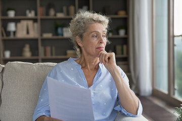 Making important decision. Pensive doubtful senior woman retiree sit on sofa hold business finance...