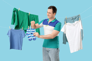 Handsome young man hanging clean clothes with wooden clothespins on blue background