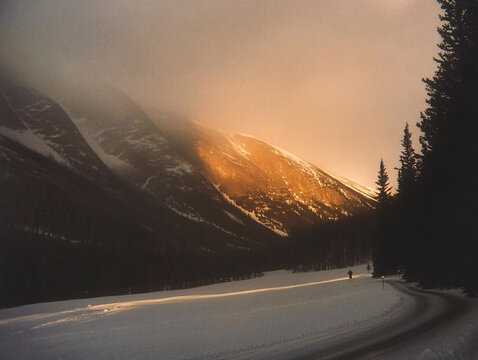 a snowy landscape at dusk or dawn, with the sun low on the horizon, casting a warm glow. A car with its headlights on is parked near a line of pine trees