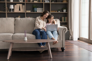 Obraz premium Weekend online at cozy home. Caring single mother embrace little daughter child sitting on couch at modern living room interior make videocall on notebook play video game learn school subject in app