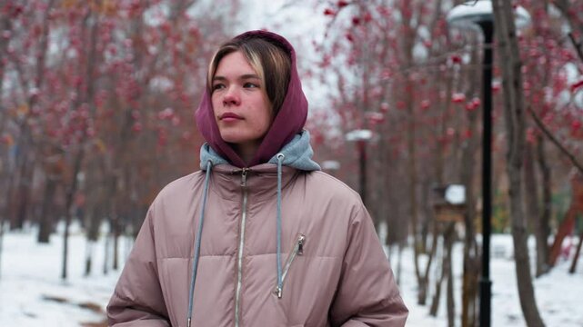 Young woman in warm hooded jacket walks along snow-covered path lined with red-berried trees, looking around contemplatively, distant blurred figure in background
