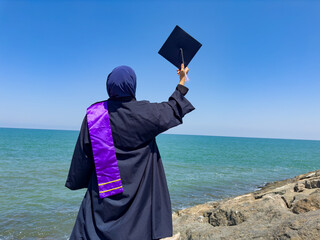 Muslim woman in graduation gown holding a mortarboard against a blue sky and sea background.  Back...