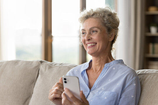 Relaxed and dreamy. Portrait of happy mindful older adult woman pensioner looking aside from smartphone screen with smile create new ideas to buy order online inspired by good news from text message