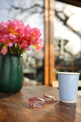 A green vase holds vibrant pink flowers beside a light blue cup on a wooden table. Rose-tinted glasses rest nearby, suggesting a serene morning atmosphere.