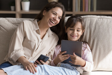 Sincere laughter and digital connection. Joyful grownup elder sister and younger one little kid girl laugh together sitting on sofa compare funny photos on pad and phone exchange memes on social media
