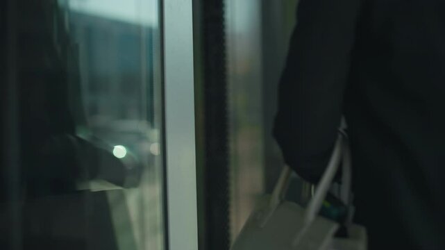 Close up of professional woman with handbag opening glass door to enter building, focus on hand gripping metal handle, reflections on door surface with urban office environment