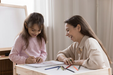 Joyful young mom older sister nanny sitting at desk laughing looking at small child girl drawing...