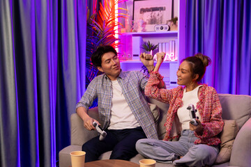 Couple gamer raising fist up holding joysticks be winner together complete level sitting sofa playing video game with front snack and drinks at neon light color living room at modern home. Infobahn. © InfiniteFlow
