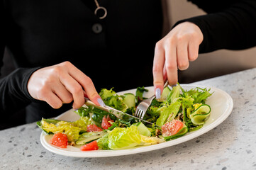 A close-up of hands gracefully cutting a fresh salad on a white plate. The salad includes various greens, cucumbers, and tomatoes, highlighting a healthy dining experience.