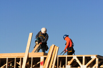 Construction workers cut and measure lumber atop a wood-framed residential structure, using power tools and tape measures during framing phase of home construction under clear morning skies