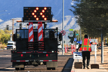 Traffic control worker places directional signage beside a barricade truck with an illuminated arrow board during lane closure setup on a city street near a school zone