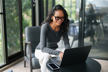 Professional Asian Businesswoman Smiling While Working On Laptop In Contemporary Glass-Walled Office Environment