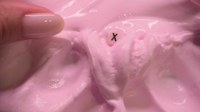 The fingers of a teenage girl are crumpling the surface of a pink slime toy that has the letter X on it.