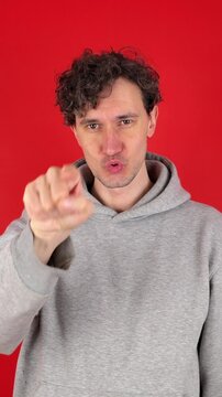 young adult curly serious man raises hand making loser gesture, L sign on forehead, teasing, accusing for defeat, expressing disrespect. Isolated red background studio shot. Concept - you lose!