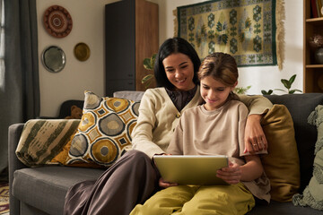 Mother and daughter smiling while using tablet together on a cozy living room sofa. Decorated in warm tones with patterned cushions and a tapestry wall hanging
