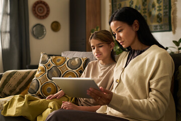 Mother and daughter reading digital content on tablet while sitting on cozy sofa in living room setting