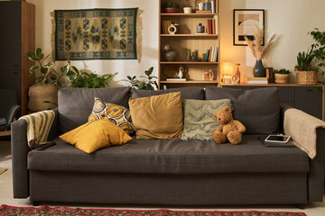 Comfortable gray sofa with various colorful pillows and brown teddy bear. Wooden bookshelves and indoor plants complement warm and inviting living room ambience