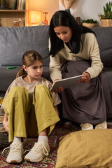 Mother and daughter engaging with tablet while sitting on living room floor. Young girl appears focused as mother provides guidance. Cozy home setting with warm lighting