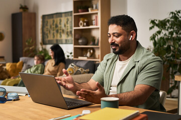 Obraz premium Man sitting in modern living room, interacting with laptop. Family in background relaxing on couch, enjoying their time together