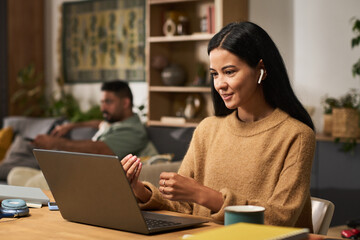 Woman smiling while wearing earbuds and working on laptop in a cozy room with another person in the background sitting on sofa