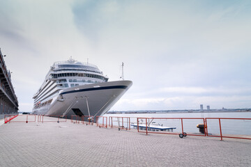 Large modern cruise ship docked at a port with a calm sea and city skyline in the background on a cloudy day.