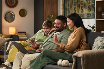 Group of three people sitting on couch, engaged with laptop, smiling and waving at screen in cozy...